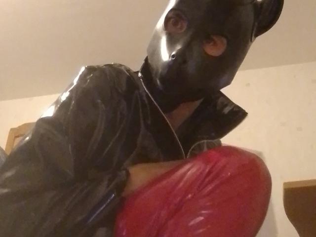 Bricebd in a black latex mask and jacket, holding red latex gloves, with visible brown eyes, in a dimly lit room. Bricebd's face is mostly covered.