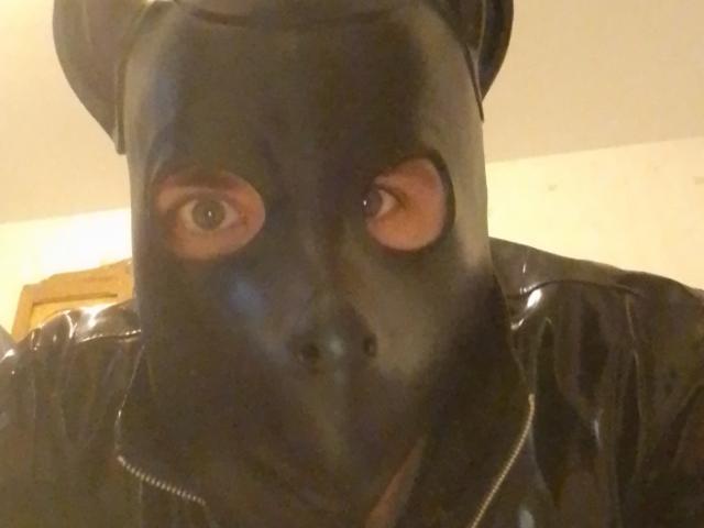 Bricebd wearing a black, shiny, full-face mask with eye holes, black leather jacket, and a black cap, looking directly at the camera with a slightly confused expression.