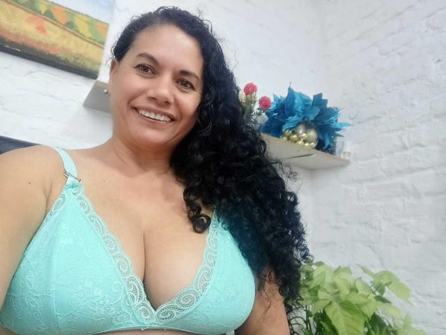 AmieLove with curly black hair, smiling, wearing a turquoise lace bra that highlights her big boobs. She's standing in a white room with a plant and blue poinsettas.