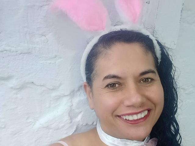AmieLove has medium brown skin, dark curly hair, and wears pink bunny ears with white fluff, a white headband, and a white choker. She's smiling with pink lipstick.