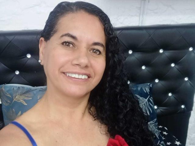 AmieLove has curly black hair, brown eyes, and a smile showing white teeth. She's wearing a blue strap, and has a red garment. Black tufted headboard with buttons.
