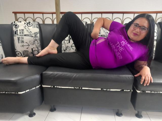 KhloeThomson, with black hair, glasses, purple shirt, and black pants, is lying on a black leather couch, legs up, showing bare feet, and a small ass.