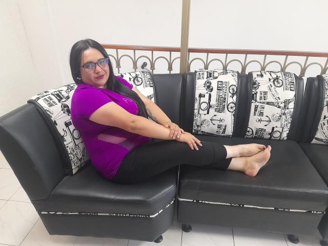 KhloeThomson with long black hair, glasses, purple top, black pants, sitting on black leather couch, legs crossed, arms on knees, bare feet, white tiled floor, black and white pillows.