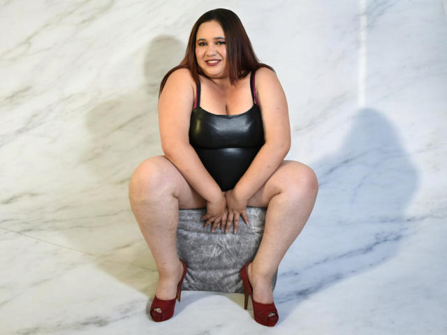KhloeThomson, chubby with pale skin, brown hair, wearing a black leather bodysuit, red high heels, squatting on a furry gray box, smiling, hands on knees.