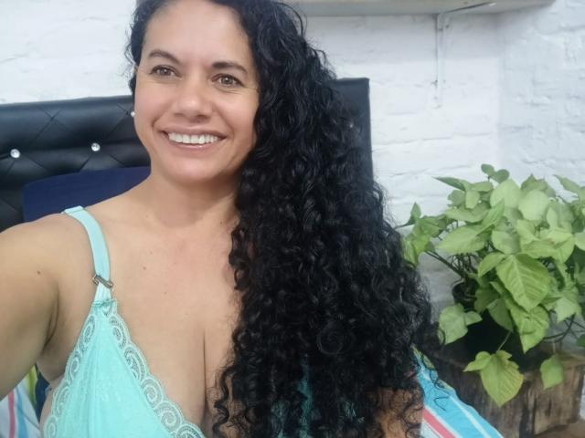AmieLove with curly black hair, smiling, wearing a light blue lace bra, showing her big tits, standing in front of a black headboard.