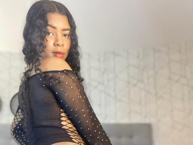 VenusSmiith with curly black hair, wearing a black mesh top with lace-up back, looking over her shoulder, red lipstick, and subtle makeup. Boobs visible through mesh.
