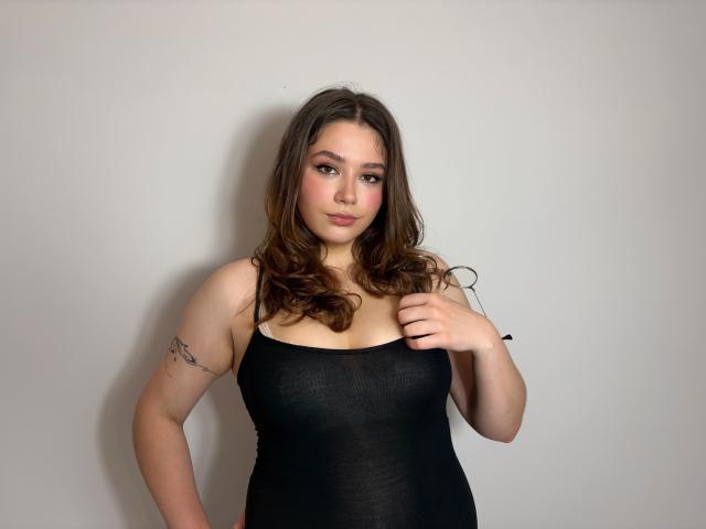 KatyAmazing69 has wavy brown hair, wearing a tight black tank top, showing her boobs and a tattoo on her right arm, standing against a plain wall.