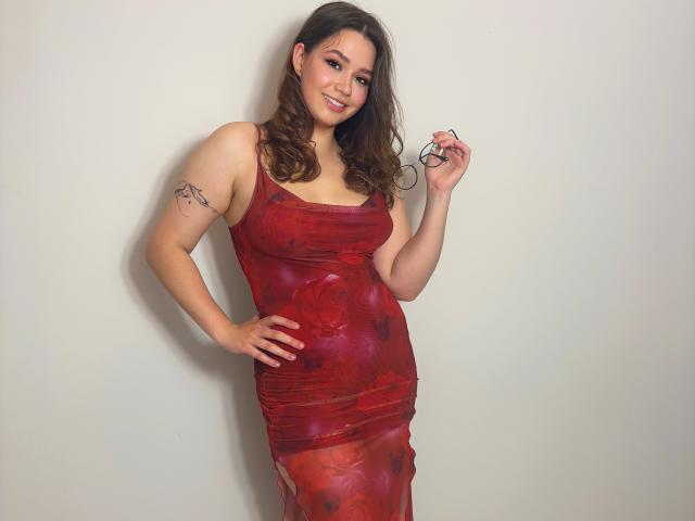 KatyAmazing69 in a red, form-fitting dress with floral patterns, holding glasses, smiling, showing a tattoo on her right arm, and standing with her hand on her hip.