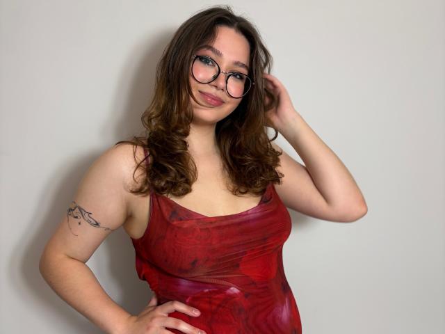 KatyAmazing69 with wavy brown hair, red tie-dye dress, glasses, and small tits, hand in hair, tattoo on right arm, smiling.