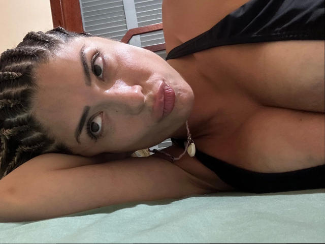 EvaBcn-hot with braided hair, pouty lips, and cleavage in a black top, lying on a green surface, head resting on arm, looking at the camera.