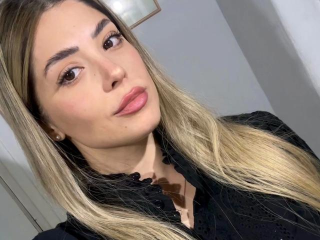 EvaBcn-hot has blonde hair, brown eyes, and pink lips. She's wearing a black shirt with a ruffled collar. Her tits are small and she's looking at the camera.