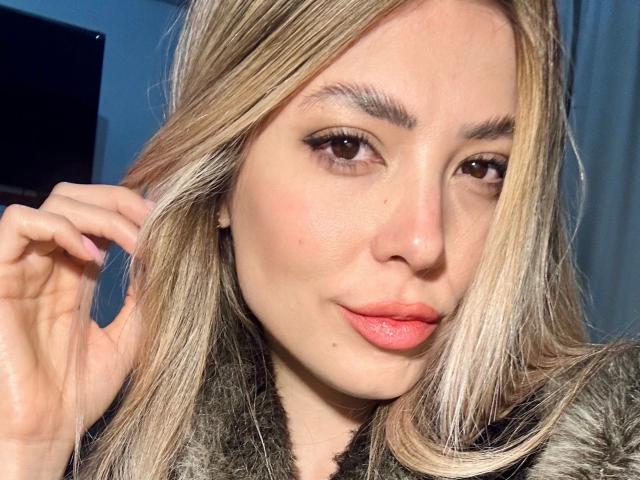 EvaBcn-hot with blonde hair, brown eyes, and pink lips, wearing a furry coat, touching her hair with a slight smile. Her tits are not visible in the image.