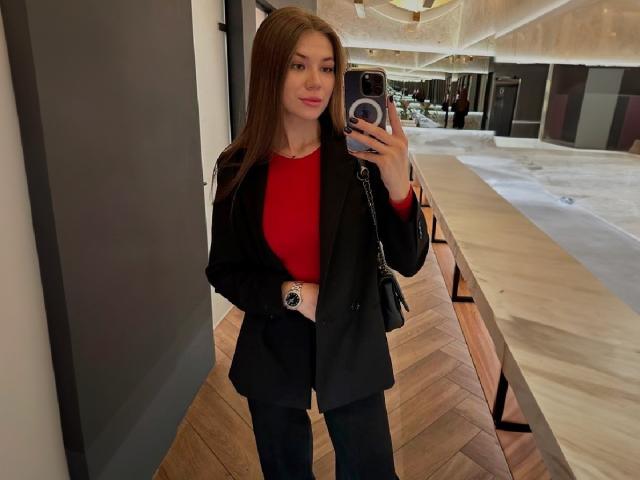 JennaCash with long brown hair, red top, black blazer, and black pants, holding a phone with a ring on it, standing in a modern, wooden hallway.