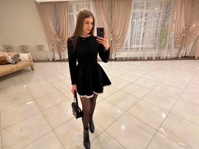 JennaCash with long brown hair, black dress, black lace trim, black stockings, black boots, holding black purse, taking mirror selfie in shiny tiled room, standing on white floor.