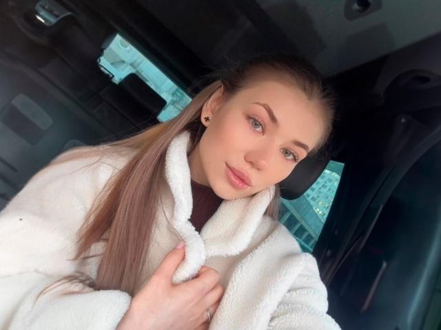 JennaCash with long brown hair, wearing a white fluffy coat, holding it close to her chest, looking at the camera with blue eyes and a subtle smile.