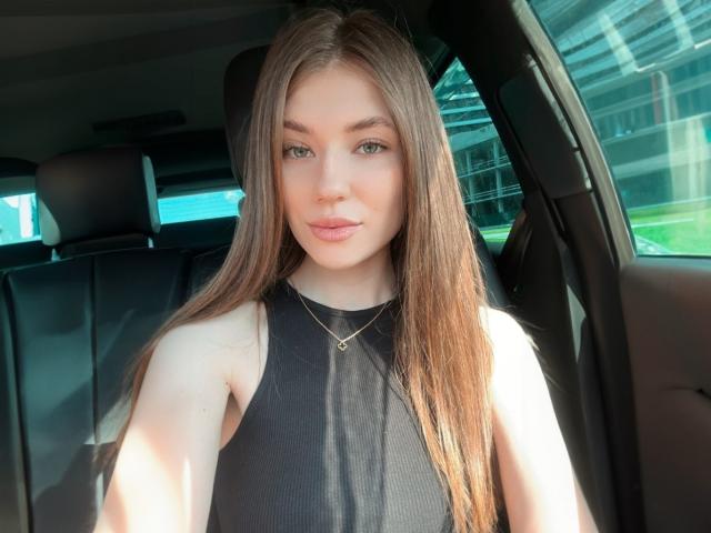 JennaCash with long brown hair, blue eyes, black sleeveless top, small nose ring, subtle makeup, slight smile, sunlight in car, wearing thin gold necklace, looking at camera.