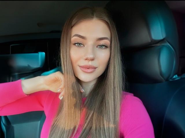JennaCash with long brown hair, green eyes, pink shirt, and subtle smile, hand in hair, sitting in car, black headrests behind her.