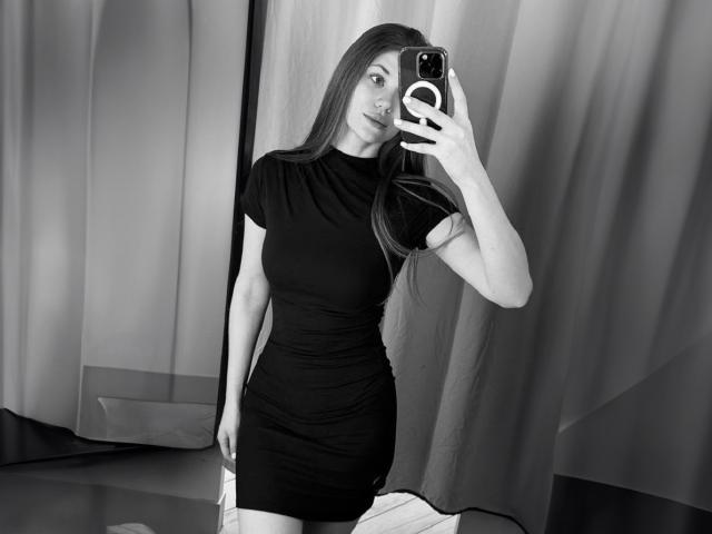 JennaCash with long straight hair, wearing a tight black dress, taking a mirror selfie with her phone, showing her small ass and medium tits, in a grayscale photo.