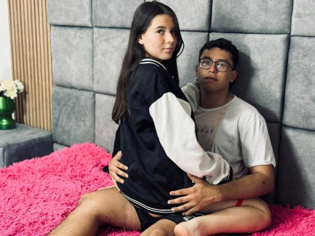 BellaThorSexUnlimit with long brown hair, wearing a black and white jacket, sits on a pink fluffy bed, legs apart, as a nerdy guy with glasses, in a white tee, grips her ass.