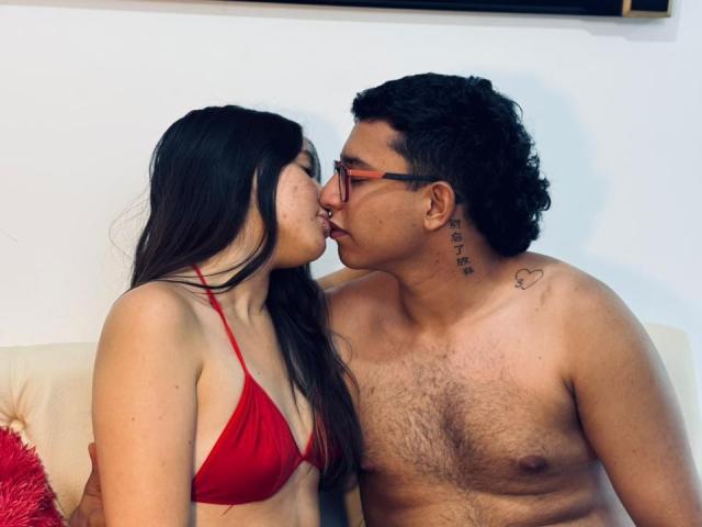 BellaThorSexUnlimit with long black hair and red bra kissing a shirtless, hairy-chested man with curly black hair and glasses. Both have nose to nose, eyes closed.