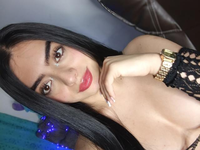 EmaRosee with long black hair, brown eyes, red glossy lips, gold watch, black fishnet sleeve, and white nails, touching her chin, showing her tits.