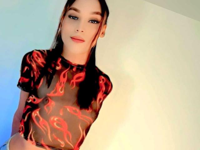 HapperTyla with long black hair, wearing a sheer black top with red flame patterns, showing small tits and a slim, sexy body, smiling slightly, with blue light on left.