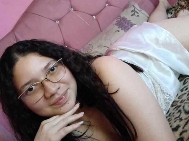 SofiSexSquirt lying on a bed, wearing glasses and a white lace dress, with long black hair, smiling, and her hand near her face, showing a small facial mole.