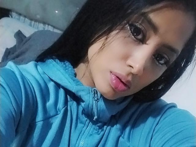 MariaCatalina with dark hair, blue hoodie, and pink lipstick, looking at the camera with slightly parted pink lips, brown eyes, and a subtle smile. Her face is centered.