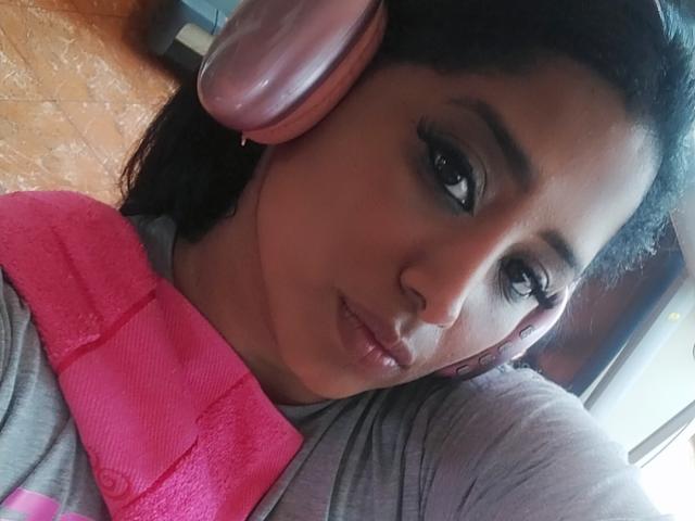 MariaCatalina with dark hair, wearing pink headphones, grey shirt, and pink towel, has pouty lips and slightly raised eyebrows, looking into camera. Her tits are not visible.