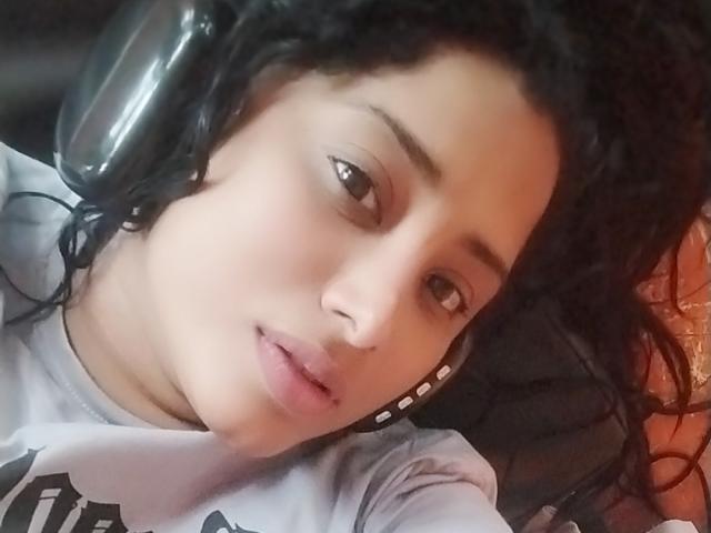 MariaCatalina with curly black hair, wearing headphones, looking at the camera with slightly parted pink lips, wearing a white shirt with black text. She has a light brown skin tone.