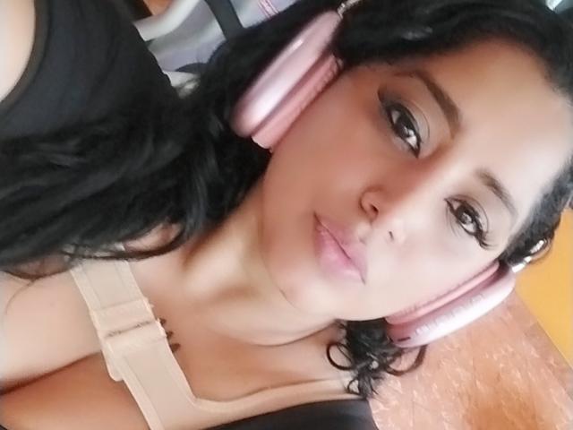 MariaCatalina with pink headphones, black wavy hair, and light skin, wearing a beige top with a low neckline, showing her tits. She has a slight smile and eyeliner.