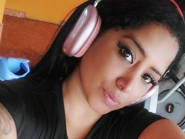 MariaCatalina with pink headphones, pouty lips, black shirt, tattoo on arm, and brown eyes, looking at camera. She's got dark hair, and a cute, sexy face.