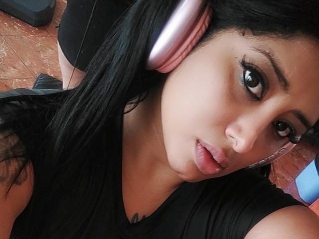 MariaCatalina with pink headphones, black hair, and brown eyes, wearing a black shirt, looking at the camera with pouty lips. Her face is in the foreground, slightly tilted.