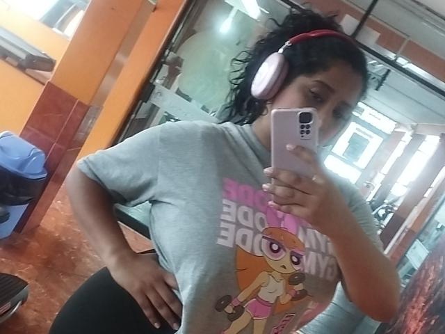 MariaCatalina with curly black hair, wearing pink headphones, a gray t-shirt with a pink cartoon character, taking a mirror selfie in a gym. Her hand's on her ass.