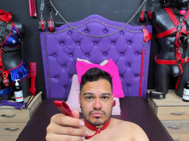 StevenHills, shirtless with a red collar, has short dark hair and a beard, holding a red dildo, with a purple tufted headboard and pink pillow behind him.