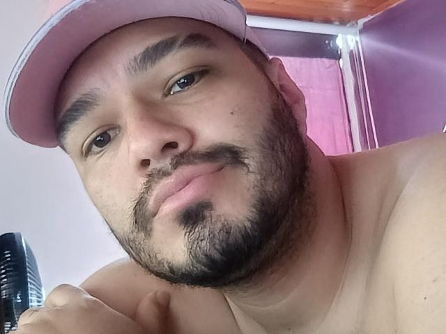 StevenHills, shirtless, wearing a pink cap, with a beard and mustache, looking at the camera, slightly smiling, holding a black fan in the bottom left.