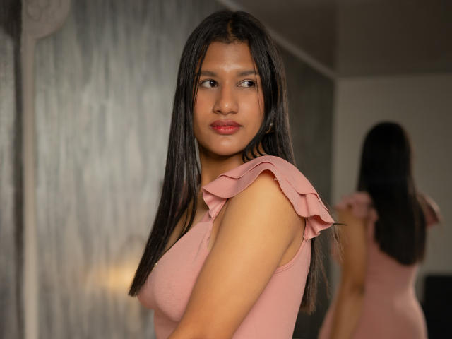 CassieJonnes with long black hair, pink ruffled sleeve top, and medium tits, looking at camera with slight smile, reflected in mirror, in soft indoor light.