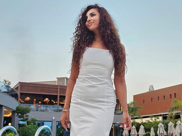 KayaOcean with long curly brown hair, white strapless dress, and a cute smile, standing outside, looking to the side, with a confident and relaxed vibe.