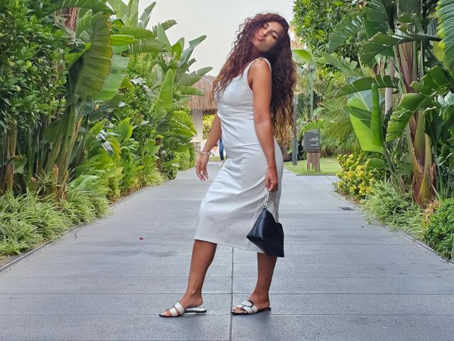 KayaOcean, avec ses cheveux bruns bouclés, sa robe blanche sans manches, son sac à main noir et ses sandales blanches, se tient sur un chemin gris, entourée de luxuriantes plantes de bananes vertes..