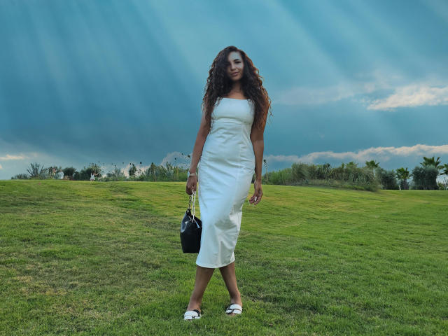 KayaOcean, com longos cabelos cacheados castanhos, vestindo um vestido branco sem alças, em pé na grama verde, segurando uma bolsa preta, usando sandálias brancas, sob um céu azul com nuvens..