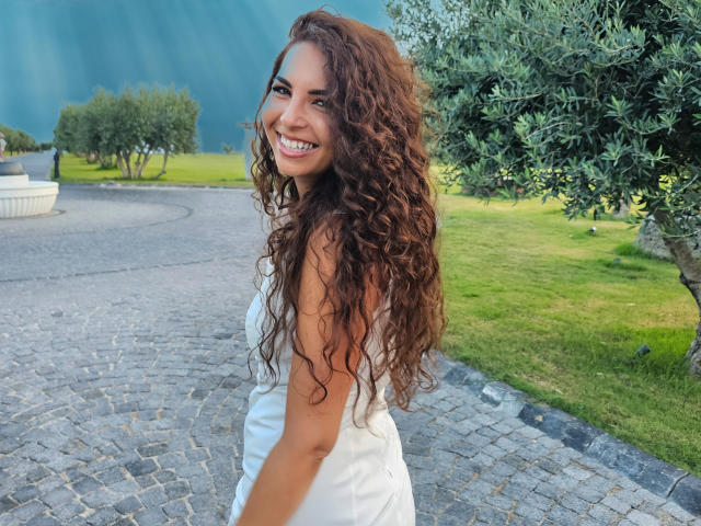KayaOcean has curly brown hair, a cute smile, and is wearing a white dress, showing her ass slightly, standing on a cobblestone path with green grass.