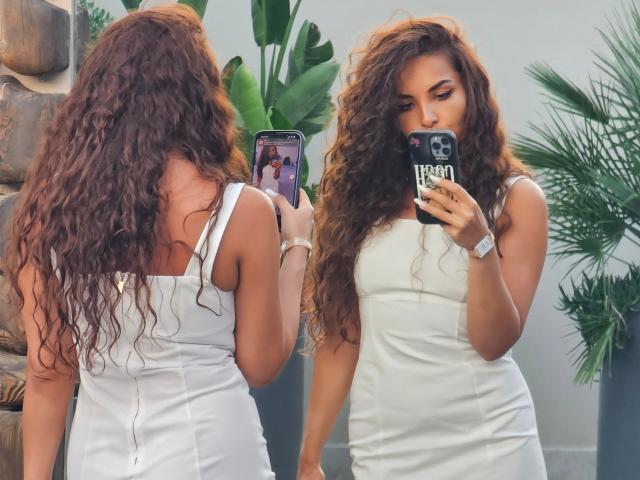 KayaOcean mit langen, lockigen, braunen Haaren, trägt ein eng anliegendes, weißes Kleid und macht ein Selfie mit ihrem Handy, wobei ihr Hintern und ihre kleinen Brüste zu sehen sind, in einem sonnendurchfluteten Raum..