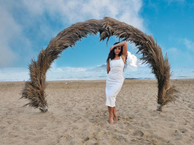 KayaOcean está descalza bajo un arco de hierba, vestida con un vestido blanco, sin tirantes y hasta los tobillos, con una mano en la cabeza, mostrando sus glúteos y pechos pequeños. Playa, cielo azul..