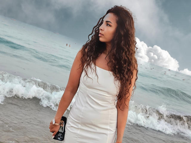 KayaOcean, con i capelli castano ricci e un abito bianco senza spalline, tiene in mano una borsa nera e si trova su una spiaggia con onde e un cielo nuvoloso. Il suo seno è di medie dimensioni..