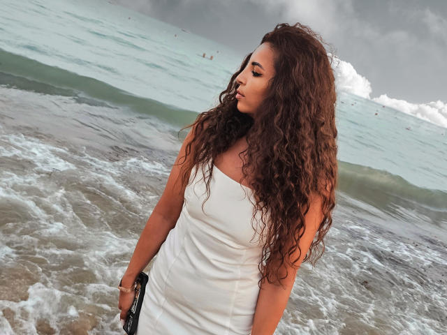 KayaOcean, con el pelo largo, rizado y castaño, vestida con un vestido blanco sin tirantes, de pie junto al océano, sosteniendo un bolso negro, mirando hacia un lado, bajo un cielo nublado..