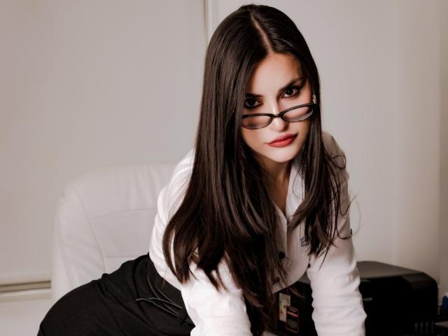 JadeAgliyah with long brown hair, glasses, white shirt, black skirt, and red lipstick, leaning forward on white leather chair, looking seductively at camera.
