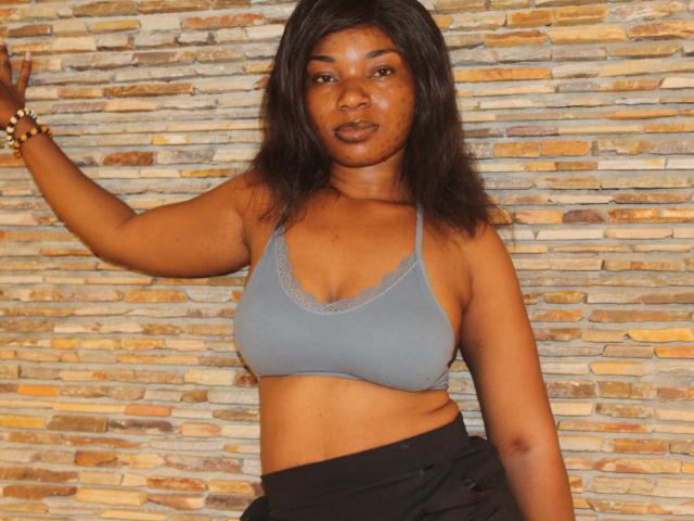 Makena69 has brown skin, black hair, small tits in a gray lace-trimmed bra, and a black skirt, standing against a stone wall, looking at the camera.