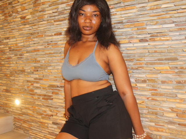 Makena69 with dark skin, medium boobs, wearing a gray sports bra and black shorts, standing against a stone wall, looking at the camera with a neutral expression.