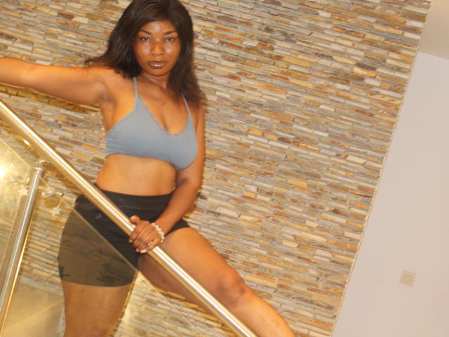 Makena69 with dark brown skin, black hair, wearing a grey sports bra and black skirt, leaning on a metal railing, showing her ass and tits, standing in a room.