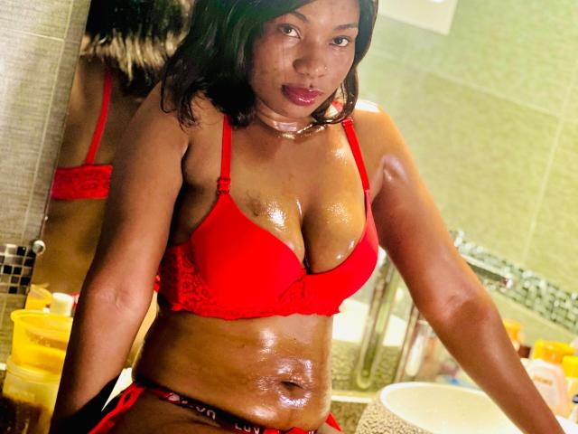 Makena69 with shiny, oiled-up brown skin, wearing red lace bra and panties, has large, glistening tits and a visible belly button, leaning forward in a bathroom.